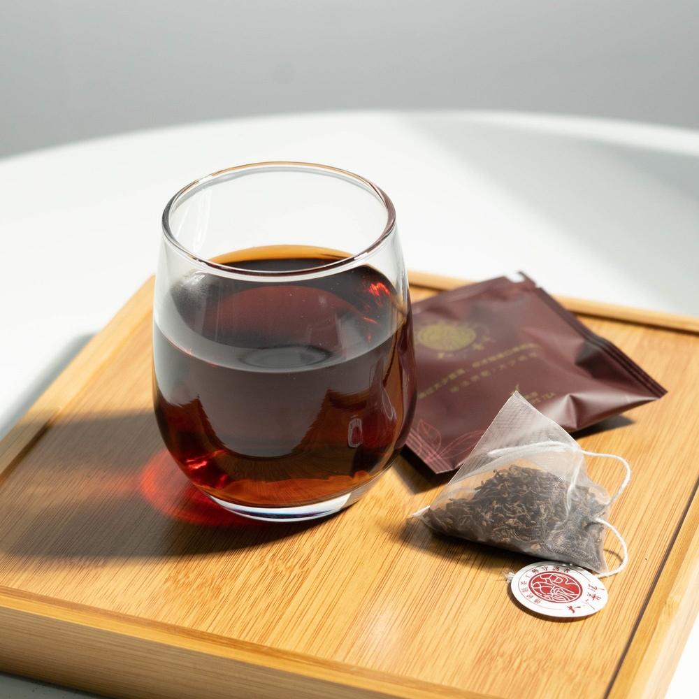 Ember Pouch triangular ripe Pu-erh Tea sachet beside its wrapper and a glass of dark tea on a wooden tray.